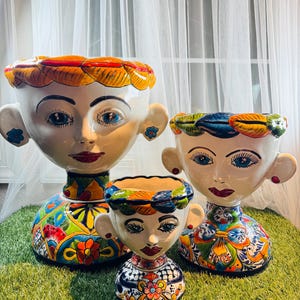 May include: Three ceramic face planters with colorful floral and geometric patterns. The planters have white faces with blue eyes, red lips, and decorative headbands. The largest planter is approximately 12 inches tall, the medium one is 9 inches, and the smallest is 6 inches.