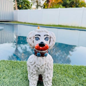 May include: A white ceramic poodle ornament with blue eyes, a red mouth, and a brown collar with blue accents. The poodle has curly fur and is standing on green grass. The background includes a swimming pool and a white fence.