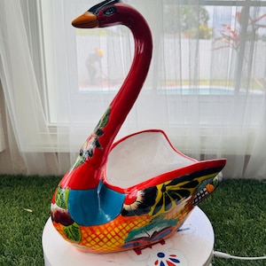 May include: A colourful, hand-painted ceramic swan planter. The swan has a red neck and head, an orange beak, and a body decorated with floral and geometric patterns. The planter has a white interior and sits on a white base.