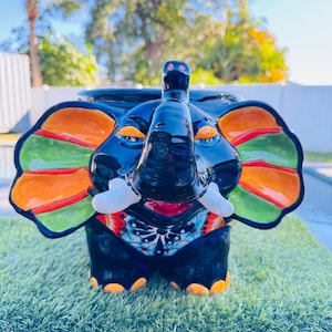 May include: A ceramic elephant figurine with a glossy black finish. The elephant has large, colorful ears with orange, green, and red accents. Its tusks are white, and the feet are orange. The figurine is decorated with a detailed, colorful pattern.