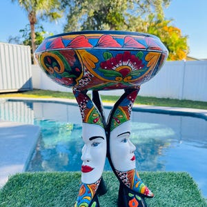May include: A decorative ceramic bowl with a colorful floral design, supported by two stylized faces with red lips and blue eyes. The bowl is painted with red, blue, yellow, and green accents. The base is black with colorful details.