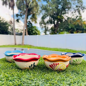 May include: Four small, decorative ceramic bowls with scalloped edges. Each bowl has a different color rim: blue, red, orange, and dark blue. The bowls are decorated with colorful floral designs and sit on a bed of green grass.