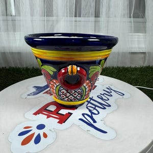 May include: A blue and yellow ceramic planter with a red ring handle and floral designs. The planter is sitting on a white surface with a sticker that says "Rio Pottery".