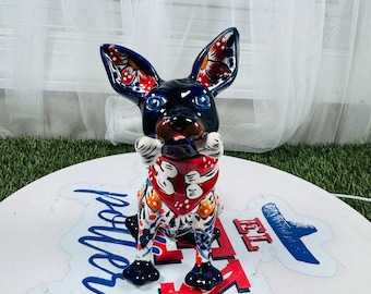 Könnte beinhalten: Keramikhundefigur mit schwarzem Körper, großen Ohren mit floralem Design und blauen Augen. Er trägt ein rotes Halstuch und hält einen Knochen. Die Figur steht auf einer weißen Oberfläche mit blauem und rotem Text.