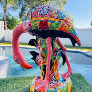 May include: A brightly coloured ceramic flamingo sculpture. The flamingo has a pink body, a long curved neck, and a patterned design on its head and base, with red, blue, green, and yellow accents. The sculpture is placed on a patch of green grass.