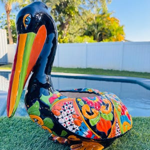May include: A ceramic pelican planter with a black body and vibrant, colorful floral patterns. The pelican has a long, multi-colored beak and is perched on green grass. The planter is designed for outdoor use.