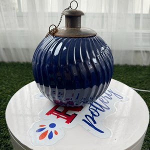 May include: A round, navy blue ceramic lantern with a ribbed design. The lantern has a metal top and a chain for hanging. The lantern is sitting on a white surface with a sticker that says "Pottery".