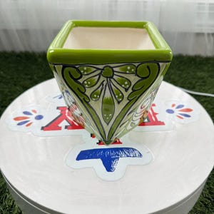 May include: A green ceramic planter with white and green floral designs. The planter has a square shape with a slightly flared top.