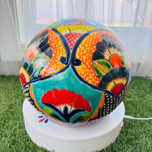 May include: A hand-painted, round ceramic decorative bowl. The bowl features a vibrant floral design with red, orange, green, and blue accents. The bowl is resting on a white rotating stand, set against a backdrop of green grass.