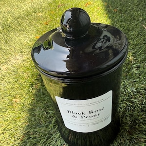 May include: A glossy black jar with a matching lid and a spherical knob. The jar has a white label with the text "Black Rose & Peony". The jar is sitting on green grass.
