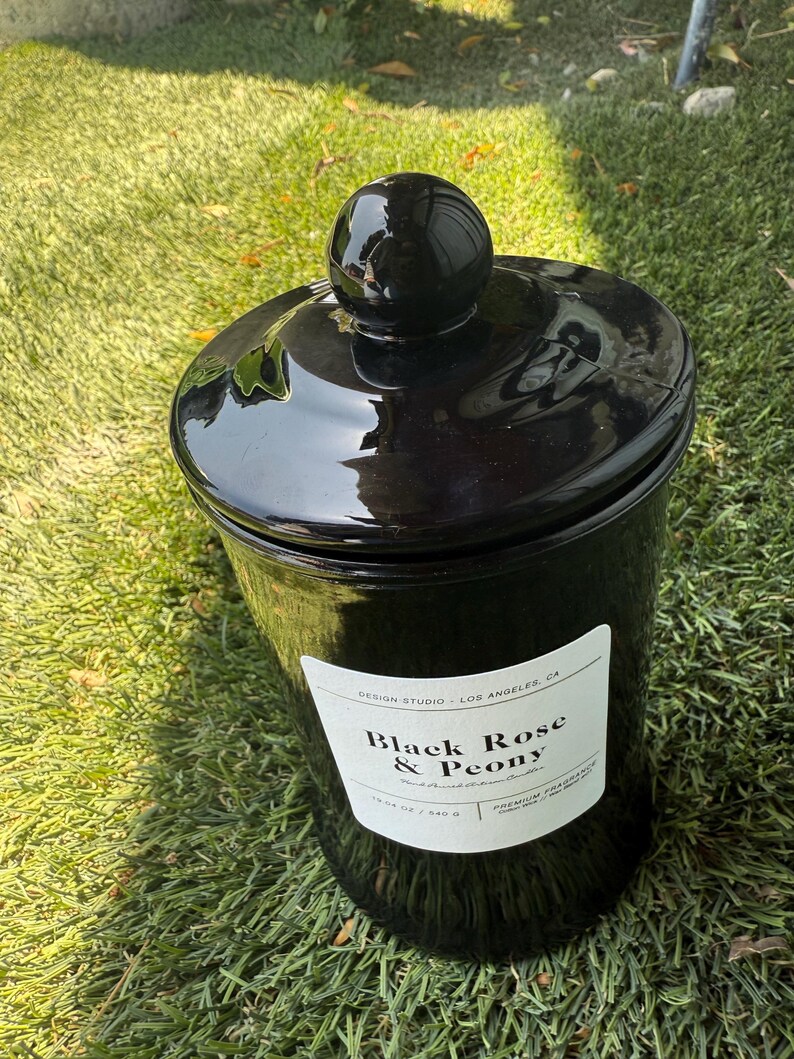 May include: A glossy black jar with a matching lid and a spherical knob. The jar has a white label with the text "Black Rose & Peony". The jar is sitting on green grass.