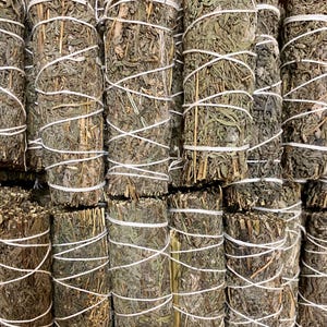 May include: A close-up of a bundle of dried sage smudge sticks. The sticks are tied together with white string and are arranged in a neat stack. The sage is a light green colour and has a slightly brown hue.