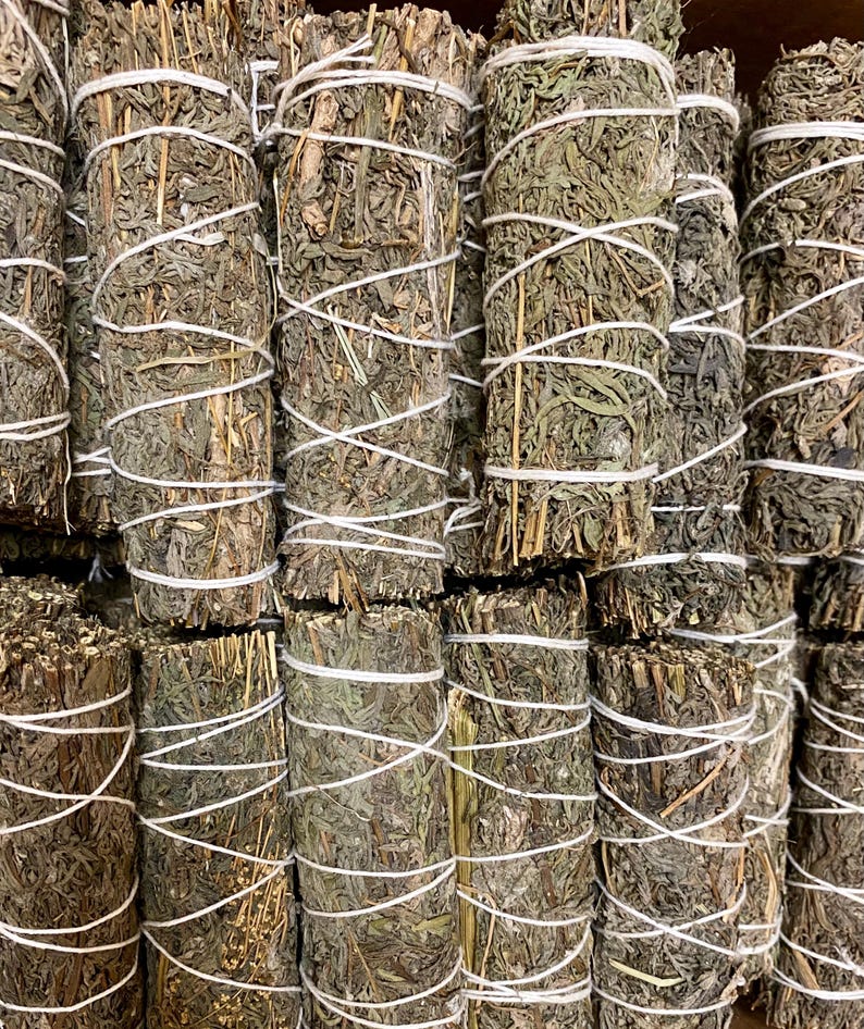 May include: A close-up of a bundle of dried sage smudge sticks. The sticks are tied together with white string and are arranged in a neat stack. The sage is a light green colour and has a slightly brown hue.