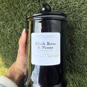 May include: A glossy black candle jar with a matching lid. The label reads "Black Rose & Peony" and "Hand Poured Artisan Candles." The jar contains 19.04 oz / 540 g of wax. The candle sits on a bed of green grass.