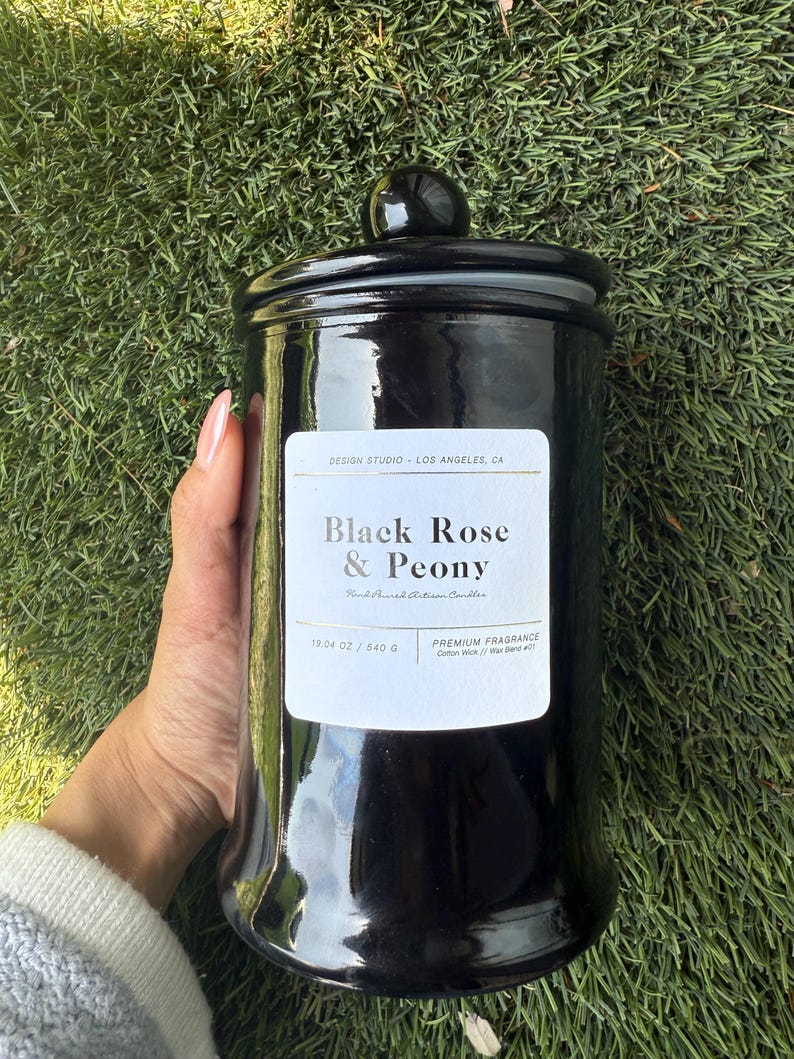 May include: A glossy black candle jar with a matching lid. The label reads "Black Rose & Peony" and "Hand Poured Artisan Candles." The jar contains 19.04 oz / 540 g of wax. The candle sits on a bed of green grass.