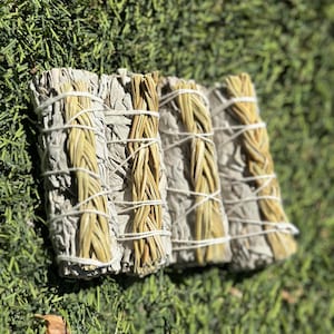 Puede incluir: Tres varitas de salvia blanca envueltas en hierba seca trenzada. Las varitas están atadas con cuerda y están dispuestas en fila sobre una superficie verde cubierta de hierba.