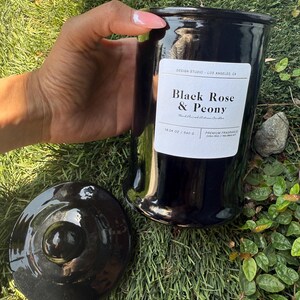 May include: A black ceramic candle jar with a matching lid. The jar has a label that reads "Black Rose & Peony" and "19.04 oz / 540 g". The candle is a premium fragrance and is hand-poured. The jar is held by a person.