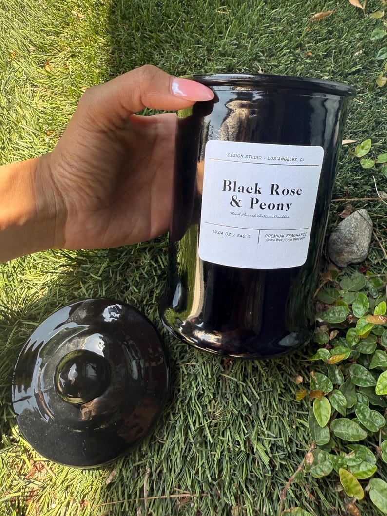 May include: A black ceramic candle jar with a matching lid. The jar has a label that reads "Black Rose & Peony" and "19.04 oz / 540 g". The candle is a premium fragrance and is hand-poured. The jar is held by a person.