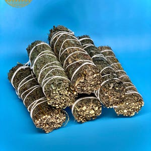 May include: A close-up of a bundle of dried sage smudge sticks tied with twine. The sticks are a dark green colour and have a rough texture. They are arranged in a pile on a bright blue background.