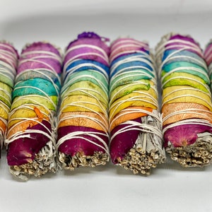 Peut inclure: Six bâtonnets de sauge arc-en-ciel liés avec de la ficelle blanche et décorés de pétales de fleurs séchées. Les bâtonnets de sauge sont disposés en rangée sur une surface blanche.