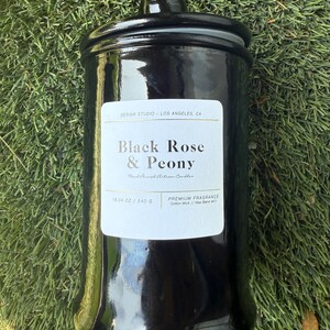 May include: A black, glossy candle jar with a matching lid. The label reads "Black Rose & Peony" and "Hand-Poured Artisan Candles." The jar contains 19.04 oz of wax. The candle sits on a bed of green grass.