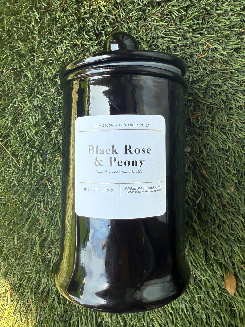 May include: A black, glossy candle jar with a matching lid. The label reads "Black Rose & Peony" and "Hand-Poured Artisan Candles." The jar contains 19.04 oz of wax. The candle sits on a bed of green grass.