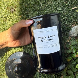 May include: A black candle jar with a lid, labeled "Black Rose & Peony." The jar is 19.04 oz and 540 g. The candle sits on a bed of green grass. The label also reads "DESIGN STUDIO - LOS ANGELES, CA."