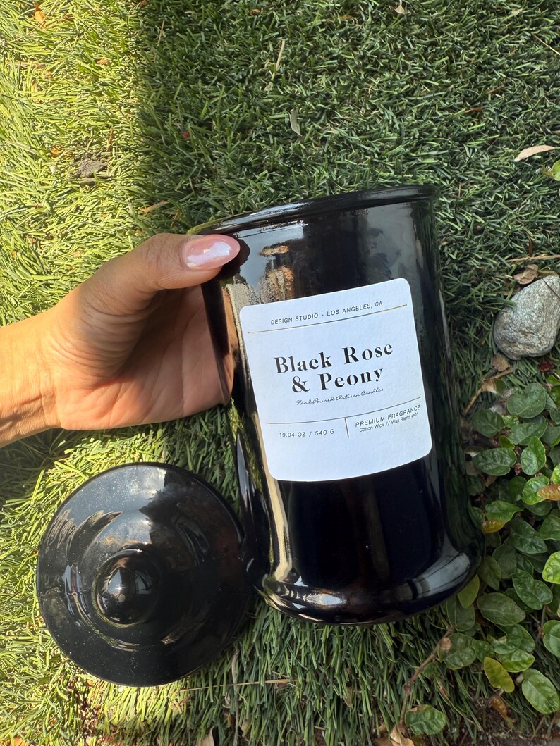 May include: A black candle jar with a lid, labeled "Black Rose & Peony." The jar is 19.04 oz and 540 g. The candle sits on a bed of green grass. The label also reads "DESIGN STUDIO - LOS ANGELES, CA."