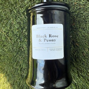 May include: A glossy black candle jar with a matching lid, set against a green grassy background. The label reads "Black Rose & Peony" and "19.04 oz / 540 g". The candle is a premium fragrance with a cotton wick.
