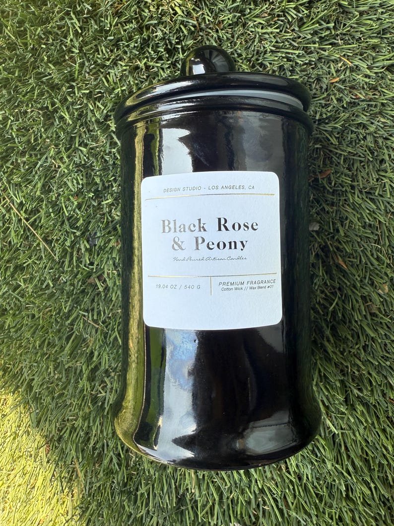May include: A glossy black candle jar with a matching lid, set against a green grassy background. The label reads "Black Rose & Peony" and "19.04 oz / 540 g". The candle is a premium fragrance with a cotton wick.