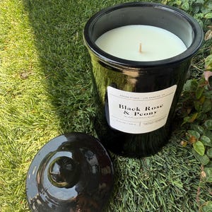 May include: A black candle in a glossy jar with a lid, sitting on grass. The candle has a white wax and a wooden wick. The label reads "Black Rose & Peony" and "Design Studio - Los Angeles, CA."