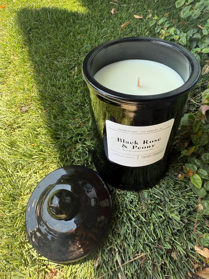 May include: A black candle in a glossy jar with a lid, sitting on grass. The candle has a white wax and a wooden wick. The label reads "Black Rose & Peony" and "Design Studio - Los Angeles, CA."