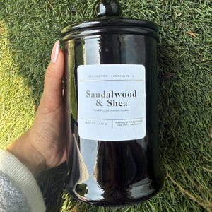May include: A black, glossy jar candle with a matching lid. The label reads "Sandalwood & Shea" and "19.04 oz / 540 g." The candle is a premium fragrance with a cotton wick and wax blend. The jar is held against a background of green grass.
