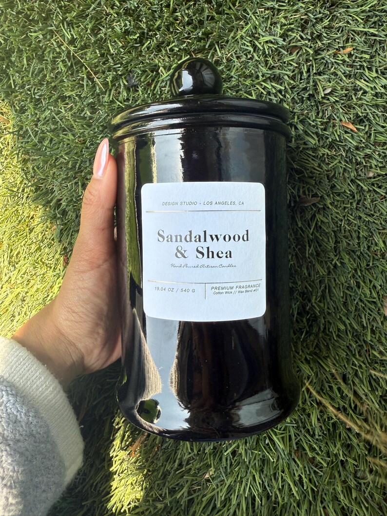 May include: A black, glossy jar candle with a matching lid. The label reads "Sandalwood & Shea" and "19.04 oz / 540 g." The candle is a premium fragrance with a cotton wick and wax blend. The jar is held against a background of green grass.