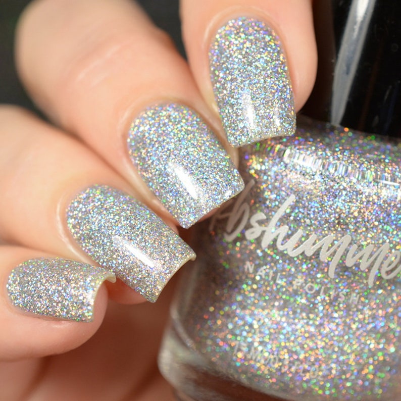 May include: A clear glass bottle of silver glitter nail polish with the brand name "Abshimmer" printed on the label. The bottle is sitting on a table with a hand wearing the nail polish.