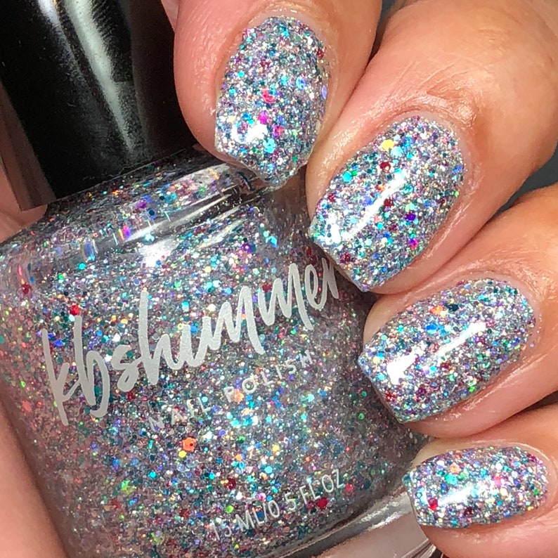 Puede incluir: Una botella de vidrio transparente de esmalte de u&ntilde;as con un acabado de brillo plateado. La botella est&aacute; etiquetada como "kb shimmer" y "nail polish". La botella se muestra con una mano que lleva el esmalte en cuatro u&ntilde;as.