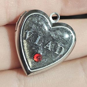 K&ouml;nnte beinhalten: Ein silberfarbenes herzf&ouml;rmiges Medaillon mit einer dunklen, glitzernden schwarzen Einlage, auf der der Name VLAD in silbernen Gro&szlig;buchstaben steht. Ein kleiner, runder, leuchtend roter Edelstein ist im unteren Teil des Herzens eingefasst.