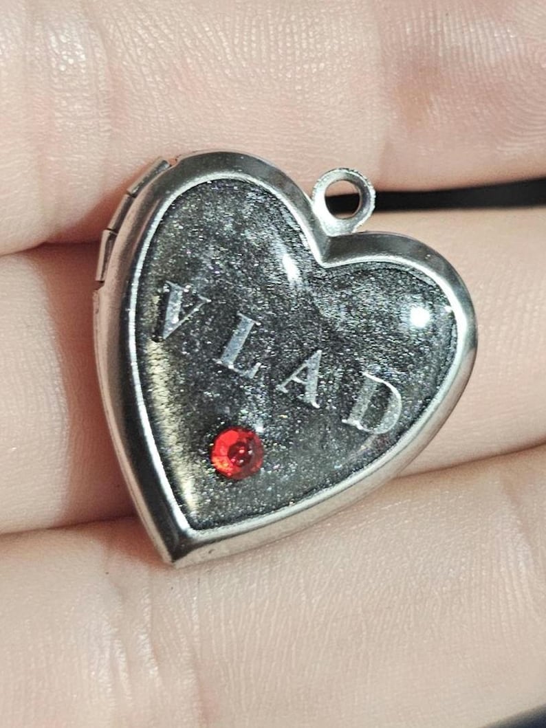 K&ouml;nnte beinhalten: Ein silberfarbenes herzf&ouml;rmiges Medaillon mit einer dunklen, glitzernden schwarzen Einlage, auf der der Name VLAD in silbernen Gro&szlig;buchstaben steht. Ein kleiner, runder, leuchtend roter Edelstein ist im unteren Teil des Herzens eingefasst.