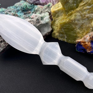 May include: White selenite crystal wand with a pointed tip and a rounded handle. The wand is carved in a unique shape and is resting on a black surface.