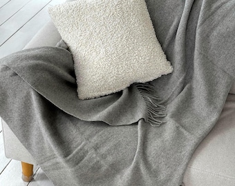 Puede incluir: Una manta gris con flecos sobre un sofá gris claro. Una almohada blanca y texturizada descansa sobre la manta. La manta y la almohada crean una estética acogedora y de tonos neutros.