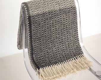 Puede incluir: Una manta con estampado de chevrón gris y blanco con flecos color crema está drapeada sobre una silla de acrílico transparente. El diseño de la manta presenta un patrón de ondas repetidas. La franja cuelga en la parte inferior.