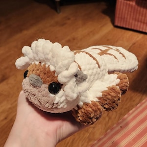 May include: A plush crochet creature featuring a white and brown color scheme. The figure has a rounded head with a white tuft of hair, large black eyes, small grey horns, and a flat tail. The body is crafted from soft, textured yarn.