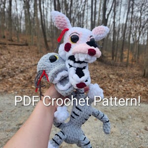 Puede incluir: Un peluche de ganchillo hecho a mano con forma de personaje con cabeza blanca, detalles rosas y detalles negros y rojos. El cuerpo es gris con rayas negras. El texto "PDF Crochet Pattern!" es visible.