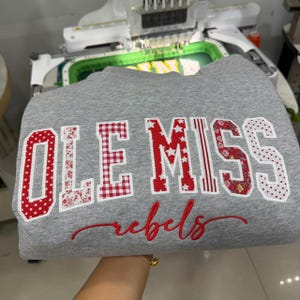 May include: A heather gray sweatshirt featuring the text OLE MISS in large, block-style letters with varied red and white patterns, including polka dots, gingham, stars, and floral prints. Below, the word rebels is embroidered in red cursive script.