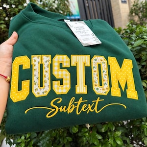 May include: A dark green crewneck sweatshirt featuring the word CUSTOM in large yellow block letters with varied patterns including polka dots, floral prints, and stripes. Below the block letters, the word Subtext is embroidered in yellow script.