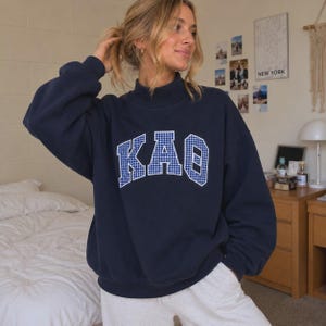 May include: A navy blue long-sleeve sweatshirt featuring a mock neck collar and the Greek letters KAO printed across the chest in a white and blue gingham check pattern. The garment has a relaxed fit with ribbed cuffs and a ribbed hemline.