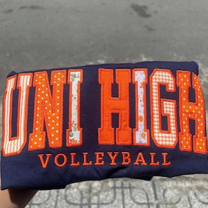 May include: A navy blue sweatshirt featuring the text UNI HIGH in large, appliqued block letters with varied orange patterns including polka dots, stripes, floral prints, and gingham. The word VOLLEYBALL is embroidered in orange below the text.