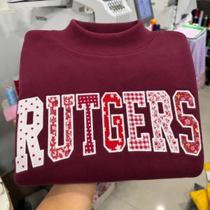May include: A maroon mock neck sweatshirt featuring the word RUTGERS in large, block-style applique letters. Each letter is crafted from a different fabric pattern, including red and white polka dots, floral prints, stars, and gingham checks.