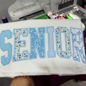 May include: A white t-shirt featuring the word SENIOR appliqued in large block letters. Each letter is outlined in light blue thread and filled with various light blue and white patterns, including floral prints, polka dots, and stripes.