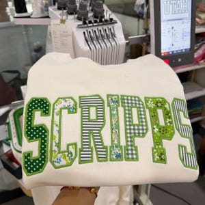 May include: A cream-colored sweatshirt featuring the word SCRIPPS in large, block-style letters. Each letter is crafted from different green and white patterned fabrics, including polka dots, floral prints, stripes, and gingham, all outlined in green.